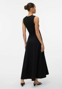 Robe maxi noire sans manches avec un corsage ajusté, une jupe évasée, une texture lisse et un design minimaliste. Portée avec des sandales noires à bout ouvert.