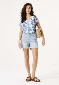 Unos pantalones cortos de mezclilla azules combinados con una camiseta de cuello en V con un estampado de flores en azul y verde. Un bolso de paja en color beige completa el conjunto.