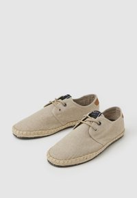 Beige Canvas-Schuhe mit Schnürdesign, Espadrille-Strohhäsole und kontrastierenden braunen Lederdetails an der Ferse.