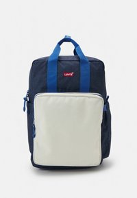 Mochila de tela azul con un bolsillo de color crema a contraste, asas superiores y cremalleras laterales. Logo rojo de Levi's en el frente. Diseño rectangular sencillo.