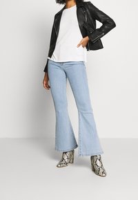 Kvinna iförd svart läderjacka, vit skjorta, ljusblå utsvängda jeans och klackboots med ormskinnsmönster mot vit bakgrund.