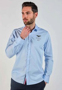 Chemise à manches longues bleu clair en tissu lisse, dotée d'un col boutonné, d'un logo brodé et d'un détail rayé contrastant au bas.