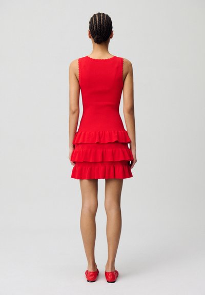 Vestito rosso senza maniche con un corpetto aderente e tre strati di volant sulla gonna. Il tessuto ha una consistenza liscia e bordi decorativi.