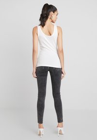 Débardeur blanc sans manches avec un dos arrondi, associé à un jean skinny gris foncé et des chaussures à talons blancs. Texture de tissu lisse.