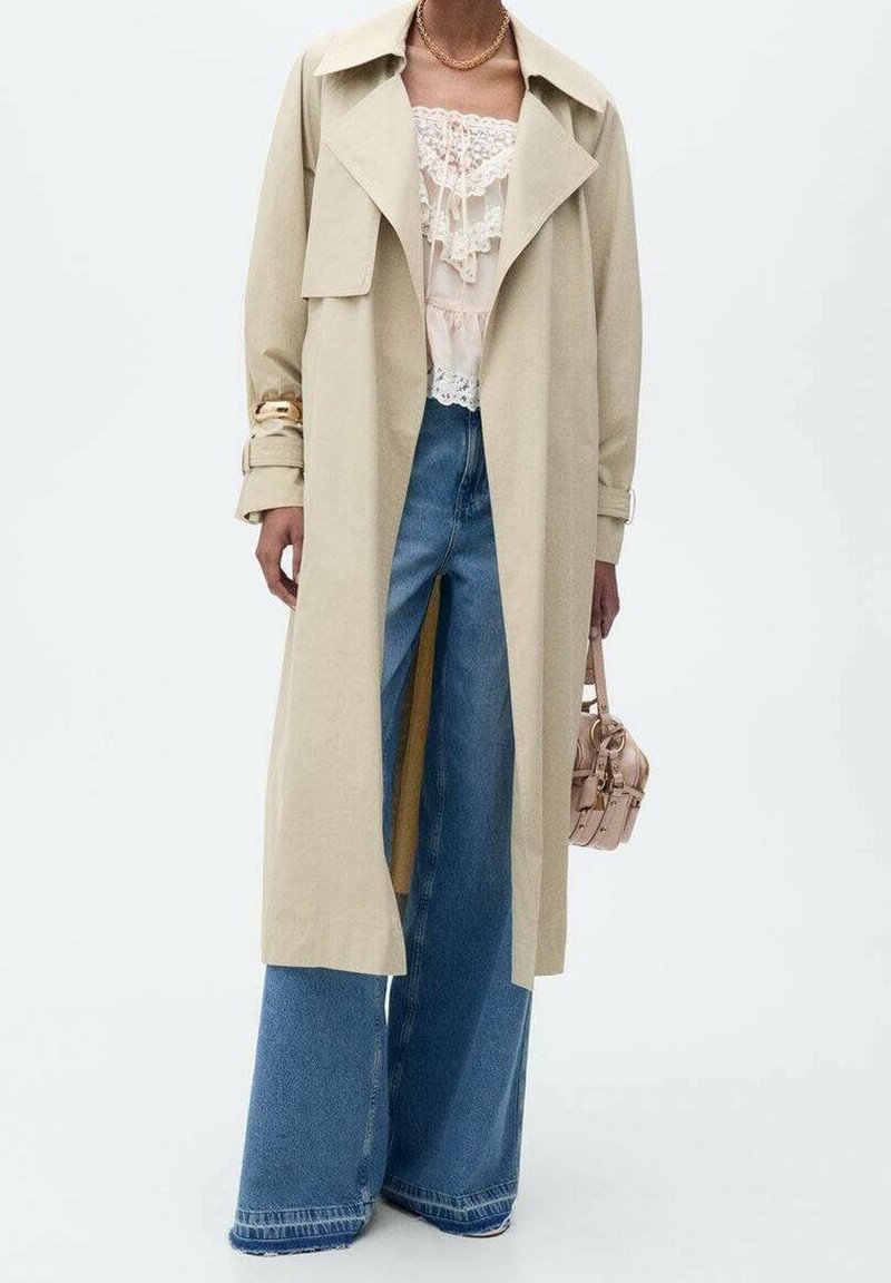 Manteau trench-coat beige par-dessus un chemisier crème orné de dentelle et un jean bleu à jambes larges, accessoirisé avec un collier en or, un bracelet et un petit sac à main beige.