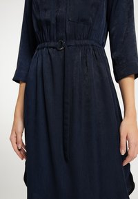 Vestido azul marino con cintura ceñida por un cinturón, con una textura suave, mangas largas y dos bolsillos en el pecho. Detalle visible de la hebilla del cinturón delgado.