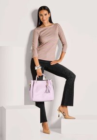 Vrouw in een beige en roze gestreepte top en zwarte jeans met rhinestones staat met één voet omhoog, terwijl ze een lavendel handtas vasthoudt en naakte pumps draagt.