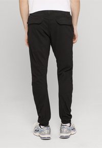 Persoană purtând pantaloni jogger cargo negri și pantofi sport albi, văzută din spate pe un fundal gri simplu.