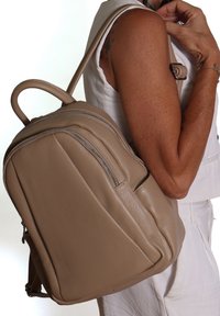 Zaino in pelle beige con forma arrotondata, dotato di due scomparti con zip e una maniglia superiore per il trasporto. Texture liscia e linee pulite.