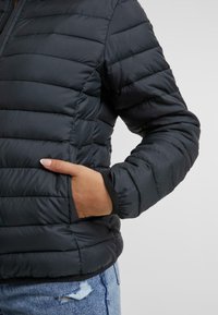 Doudoune noire avec un matelassage horizontal, dotée d'une poche latérale et de poignets côtelés. Le matériau a une apparence lisse et légèrement réfléchissante.