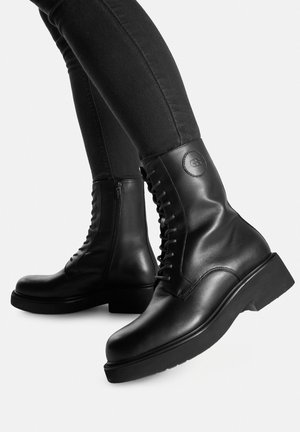 Czarne skórzane botki za kostkę z masywną podeszwą, sznurowaniem z przodu, bocznym zamkiem i okrągłą naszywką z logo na cholewce. Gładka faktura.