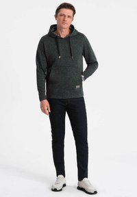Felpa con cappuccio verde scuro con un motivo texturizzato e macchiato, cordini e tasca frontale, abbinata a jeans scuri e sneakers chiare.