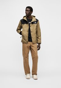 Μπεζ μπουφάν με μαύρο μπροστινό μέρος, κουκούλα και κούμπωμα με κουμπιά μπροστά. Φορεμένο πάνω από μαύρο πουκάμισο και συνδυασμένο με μπεζ παντελόνι και ανοιχτόχρωμα παπούτσια.