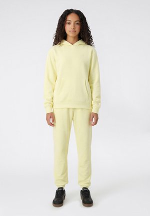 Joven con cabello rizado que lleva una sudadera con capucha amarilla pálida y pantalones deportivos a juego, de pie mirando hacia adelante sobre un fondo liso.