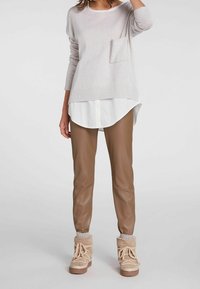 Hellgrauer Pullover mit einer Fronttasche über einem weißen Hemd mit Kragen; kombiniert mit braunen Kunstlederhosen und hellbraunen Schnürstiefeln.