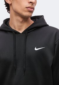 Jeune homme portant un sweat à capuche noir avec cordons, avec un petit logo Nike Swoosh blanc sur la poitrine.