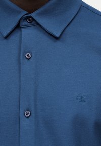 Μπλε πουκάμισο με γιακά από υφασμάτινη υφή, κούμπωμα με κουμπιά και διακριτικό κεντημένο λογότυπο "cK" στο στήθος.