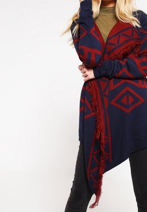 Cardigan navy con motivi geometrici rossi, frontale aperto, maniche lunghe e dettagli frangiati all'orlo. Indossato sopra una maglietta verde e pantaloni neri.