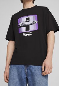 Camiseta de algodón negra con un gráfico en púrpura y blanco de un coche Porsche Turbo, con texto en negrita debajo. Corte relajado, mangas cortas.