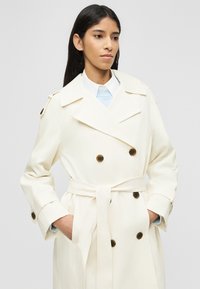 Femme aux longs cheveux foncés portant un trench-coat écru ceinturé avec des boutons foncés, debout les mains dans les poches devant un fond uni.