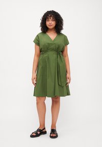 Vestito avvolgente verde con maniche corte, scollatura a V, vita con cinturino e tasche laterali, realizzato in tessuto morbido. Indossato con sandali neri.