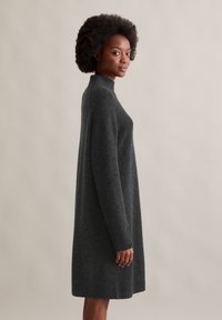 Graues, gestricktes Kleid mit hohem Kragen, langen Ärmeln und lockerer Passform. Weicher Stoff, knielang und einfarbig ohne Muster.