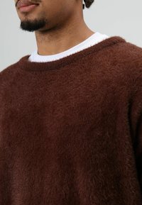 Bruine, pluizige trui met een losse pasvorm, ronde hals en zachte textuur, gedragen over een wit shirt. Close-up van de stof.