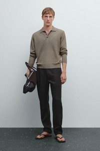 Man in een olijfgroene poloshirt met lange mouwen, zwarte broek en sandalen, die een donkerbruine leren tas naast zich vasthoudt tegen een vlakke achtergrond.