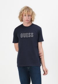 Navy blue cotton t-shirt featuring "GUESS" logo in white, short sleeves, and crew neckline. Worn with dark blue jeans.