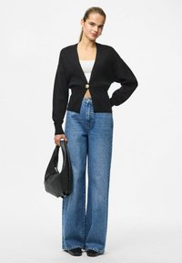 Czarny dziergany kardigan z głębokim dekoltem w serek i złotym zapięciem na guziki, zestawiony z jasnoniebieskimi szerokimi spodniami typu jeans oraz teksturowaną czarną torebką.