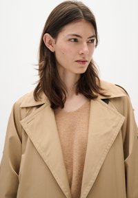 Beige trench coat with a large collar over a light brown sweater. Smooth fabric, loose fit, minimalistic style, and no visible patterns.
