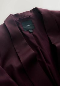Giacca stile kimono color bordeaux con una texture liscia simile alla seta. Presenta un colletto a rever e un'etichetta con la scritta "NEXT".