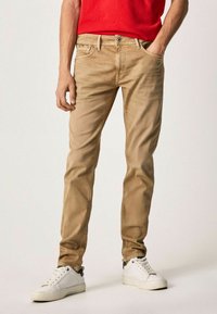 Vaqueros de algodón beige con ajuste slim, diseño de cinco bolsillos y costuras en contraste, combinados con zapatillas blancas y una camisa roja.
