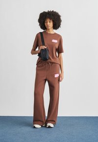 Σετ με καφέ βαμβακερό μπλουζάκι και παντελόνι φόρμας με ροζ λογότυπα. Το μοντέλο κρατά μια μικρή μαύρη τσάντα. Αθλητικά παπούτσια ολοκληρώνουν την εμφάνιση.