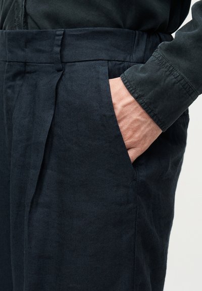 Main dans la poche d'un pantalon plissé bleu marine foncé avec une manche de chemise noire à manches longues visible.