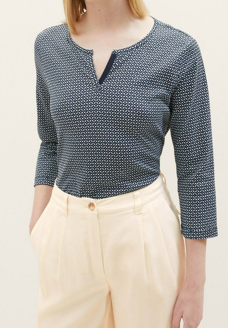 Haut à motif marine avec un décolleté en V et des manches trois-quarts, associé à un pantalon taille haute beige clair avec une fermeture à bouton unique.