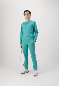 Sudadera en turquesa y joggers a juego, elaborados en algodón, con puños elásticos y bolsillos. Racket de tenis sostenido en una mano.