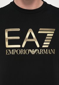 Black cotton T-shirt featuring bold gold logo "EA7 EMPORIO ARMANI" with varied font sizes and styles, round neckline, and short sleeves.