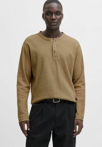 Hombre de piel oscura que lleva puesta una camiseta de mangas largas color beige y pantalones negros con un cinturón negro, de pie contra un fondo claro y sencillo.