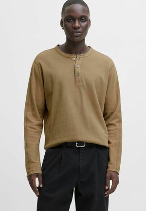 Man with dark skin wearing a tan long-sleeve henley shirt and black pants with a black belt, standing against a plain light background.