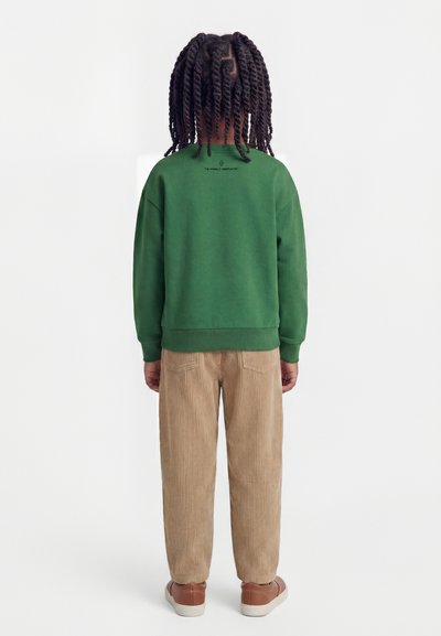 Bambino con i capelli intrecciati di spalle, indossa una felpa verde, pantaloni beige di velluto a coste e sneakers marroni su uno sfondo semplice.