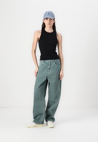 Top nero senza maniche, jeans larghi verde chiaro, sneakers bianche e un cappellino azzurro chiaro. Design semplice e casual con vestibilità rilassata.