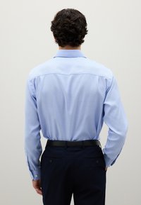 Homme aux cheveux bouclés et foncés portant une chemise à manches longues bleu clair rentrée dans un pantalon bleu marine foncé avec une ceinture noire, faisant face à un mur uni.