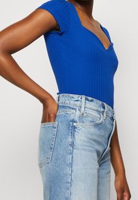 Blue ribbed bodysuit with short sleeves, V-neckline, worn with light blue high-waisted jeans featuring front pockets and a faded finish.
