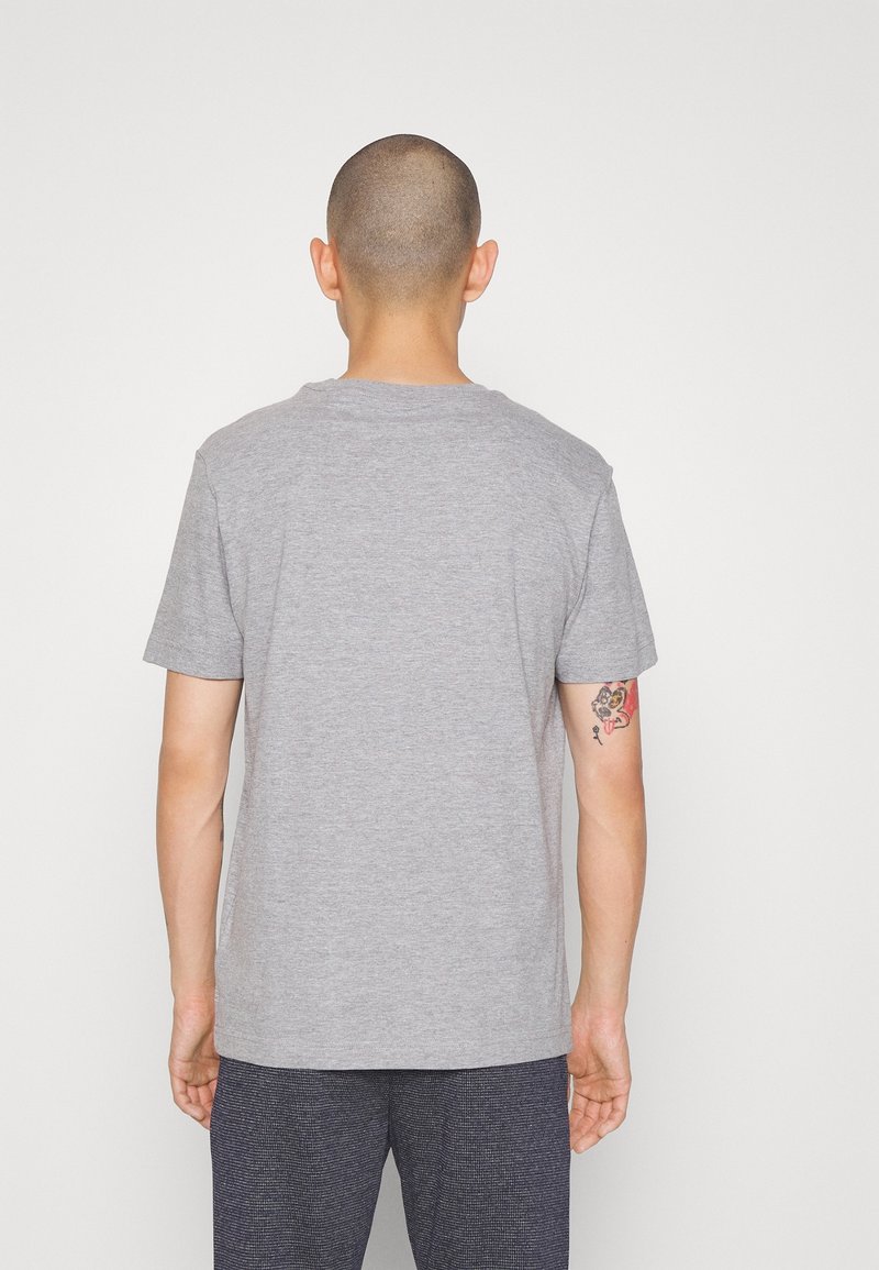 Homme avec la tête rasée portant un t-shirt gris uni et un pantalon texturé sombre, se tenant de dos face à un fond blanc.