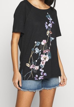 Kvinde iført en sort kortærmet skjorte med lodret blomsterprint kombineret med blå denim shorts med frynser.