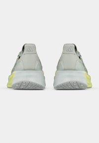 Chaussures de sport de couleur claire avec une texture lisse, dotées d'une semelle coussinée, d'accents jaunes et d'un design minimaliste, logo visible sur le talon.