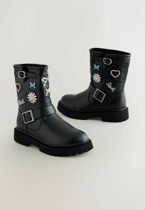 Cizme de gleznă din piele sintetică neagră, cu patch-uri decorative în formă de flori, inimioare și fluturi, o cataramă pe lateral și o talpă din cauciuc texturată.