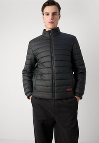 Zwarte pufferjas met een hoge kraag, gestikte horizontale lijnen, rits aan de voorkant en een rood "HUGO" logo aan de linkerkant.