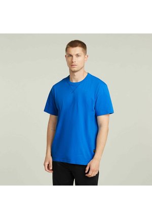 Giovane uomo con capelli corti che indossa una maglietta a maniche corte blu brillante e pantaloni neri, in piedi contro uno sfondo chiaro e uniforme.